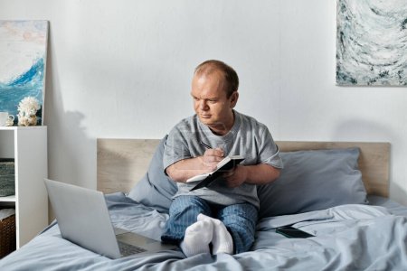 Un hombre con inclusividad se sienta en su cama, escribiendo en un cuaderno, con un ordenador portátil y teléfono a su lado.