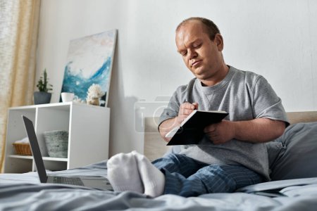 A man with inclusivity sits on his bed, writing in a notebook, with a laptop beside him.