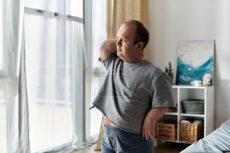 A man with inclusivity stands by a window, stretching his back in a well-lit room.