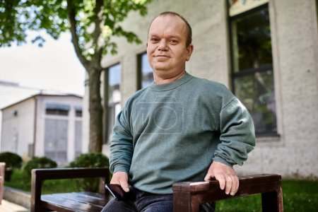 A man with inclusivity sits on a bench outdoors, looking directly at the camera.