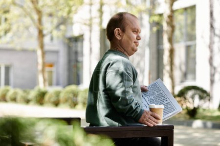 Un hombre con inclusividad se sienta en un banco en un parque, leyendo un periódico y sosteniendo una taza de café.