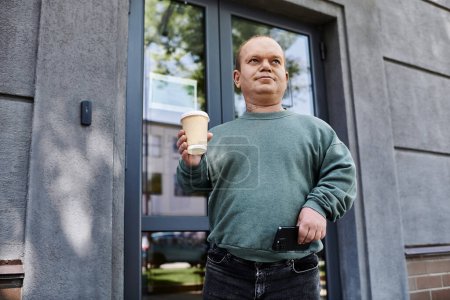 Un homme avec inclusivité se tient devant un bâtiment, tenant une tasse de café et un smartphone.