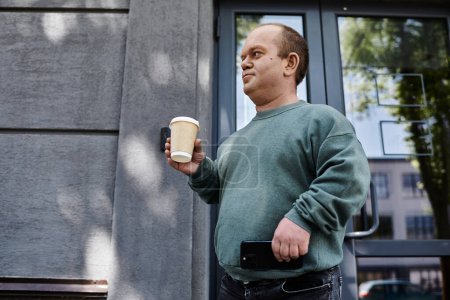 A man with inclusivity in a green sweater holds a cup of coffee while looking towards his left.