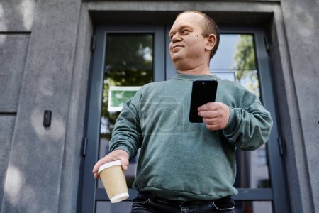 A man with inclusivity with a cup of coffee in his hand checks his phone while standing outside a building.