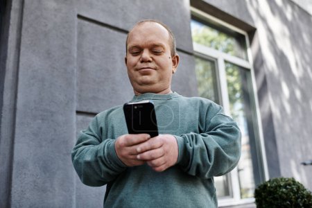 A man with inclusivity in a teal sweater checks his phone while standing on a city street.