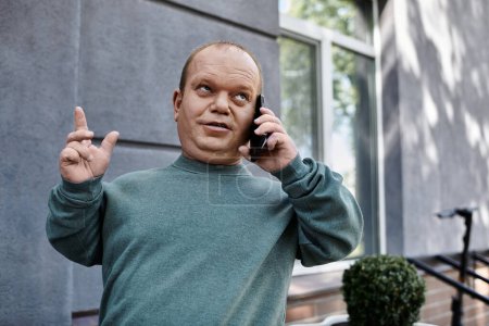 Un hombre con inclusividad vistiendo una sudadera verde se para en la calle, hablando por teléfono con un dedo levantado, posiblemente haciendo un punto o enfatizando sus palabras.
