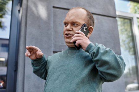 Un homme avec inclusivité dans un pull sarcelle parle animément sur son téléphone à l'extérieur d'un bâtiment.