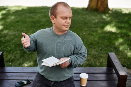 Un homme avec inclusivité est assis sur un banc de parc avec un cahier et un stylo, profitant d'une pause café.