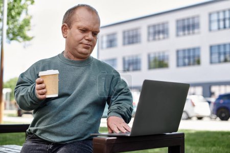 A man with inclusivity works on his laptop while enjoying a cup of coffee on a bench in a city park.
