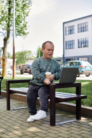 A man with inclusivity sits on a bench in a park, enjoying a cup of coffee and working on his laptop.
