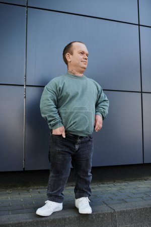 A man with inclusivity wearing a green sweatshirt and jeans stands casually by a wall.
