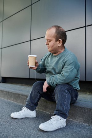 Un homme inclusif est assis sur un trottoir, tenant une tasse de café, apparemment perdu dans sa pensée.