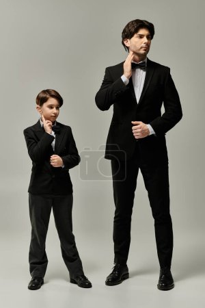 Foto de Un padre y su hijo pequeño, ambos vestidos con trajes negros, se paran uno al lado del otro, el padre ajustando su corbata. - Imagen libre de derechos