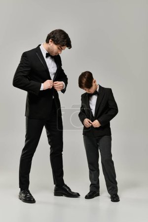 A father and son, both dressed in black tuxedos, stand together in a studio setting.