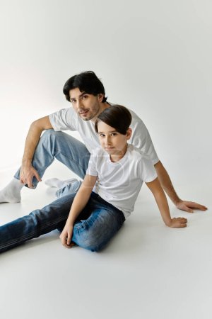 A father and his son sit on a white floor, casually dressed in white t-shirts and jeans, gazing at the camera.
