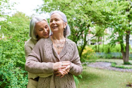 Two middle-aged women in cardigans embrace in a park, surrounded by lush greenery.