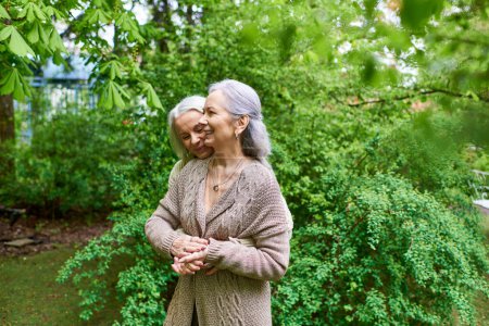 A middle-aged lesbian couple in cardigans hugs each other amidst green trees.