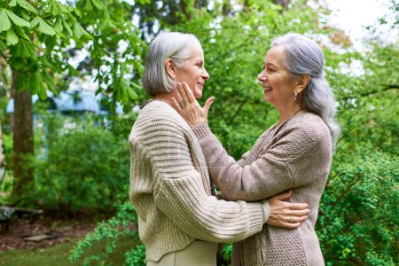 Two middle-aged women, dressed in cardigans, lovingly embrace in a lush, green garden setting.