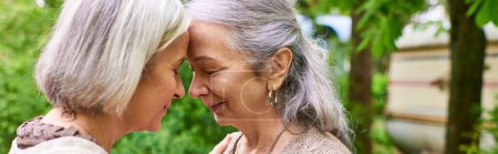 Two middle-aged women, with grey hair, stand close together and hug, eyes closed, as they share a moment of intimacy near a vibrant green tree.