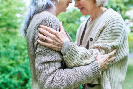A middle-aged lesbian couple embraces each other warmly while wearing cardigans, surrounded by lush green trees.