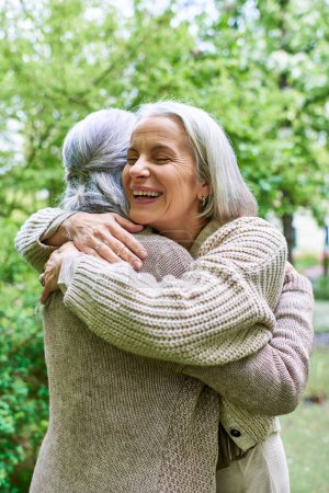 Two middle-aged women, dressed in cardigans, hug each other warmly while standing amidst a lush green backdrop.