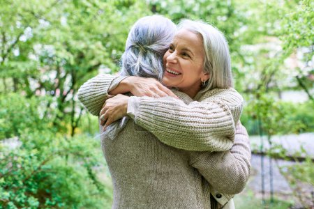 A middle-aged lesbian couple, wearing cardigans, embrace each other in a park setting.