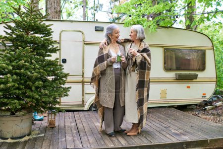 Zwei Frauen stehen in Decken gehüllt vor einem Wohnmobil und genießen einen Moment der stillen Geselligkeit in einer bewaldeten Umgebung.