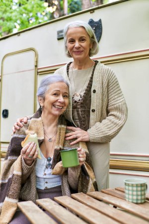 Two middle-aged lesbian women are enjoying a camping trip in a green forest. They are standing in front of their camping van.