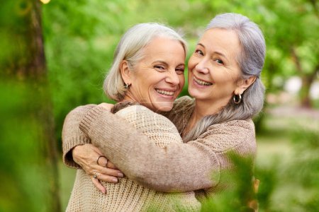 Photo for Two middle-aged women embrace in a green forest setting. They appear happy and relaxed, enjoying their time outdoors. - Royalty Free Image