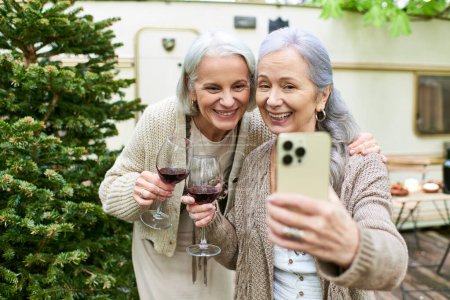 Ein lesbisches Paar mittleren Alters genießt ein Glas Wein und macht beim Zelten ein Selfie im Wald.