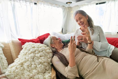 A lesbian couple, both middle-aged with gray hair, share a lighthearted moment while relaxing in a camping van.