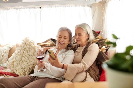Two middle-aged women relax inside a camper van, enjoying wine and using a smartphone.