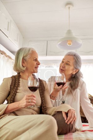 Two middle-aged women, a lesbian couple, relax in a camper van and share a toast with glasses of red wine.
