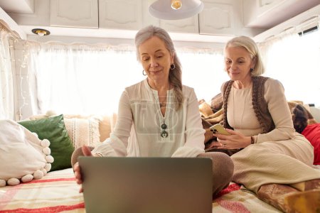 Two women sit inside a camper van, one working on a laptop, while the other sits with phone.