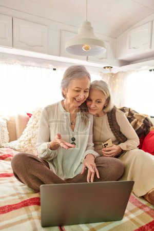 A lesbian couple smiles while using a laptop inside their camping van during a road trip.