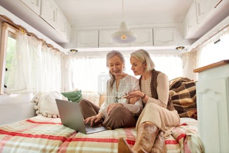 Two middle-aged women relax in a camper van, one using a laptop while the other looks on.
