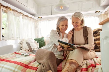 Two middle-aged women, a lesbian couple, are happily relaxing in their camper van during a trip. They are sitting on the bed, reading a magazine.