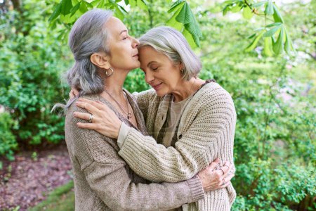 A middle-aged lesbian couple in cardigans hug each other affectionately in a lush green forest.