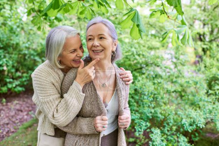 Ein lesbisches Paar mittleren Alters in Strickjacken, mit grauen Haaren, lachen und umarmen sich in einer wunderschönen grünen Gartenkulisse.