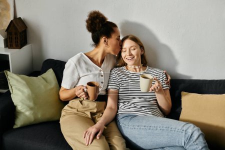 Two women, in casual wear, share a cozy moment on a couch with cups of coffee.