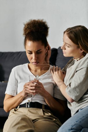 A lesbian couple shares a tender moment at home, one holding a glass of water while the other offers support and comfort.
