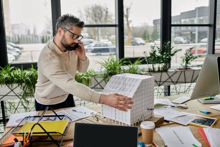 A handsome businessman with a beard works on a building model while talking on the phone in a modern office.