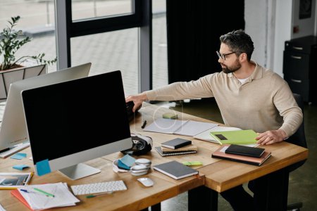 A handsome businessman with a beard works at his desk in a modern office, with two monitors and several notebooks and pens.