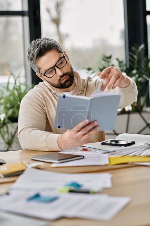 A handsome businessman with a beard is working in a modern office. He is wearing glasses and is reviewing documents on a table with a variety of other documents and office supplies.
