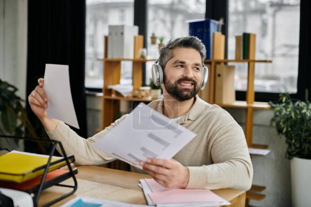 A bearded businessman with headphones on smiles while reviewing documents at his desk in a modern office setting.