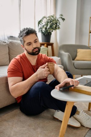 A bearded man in casual attire works remotely from home, sitting on the floor with a coffee mug in one hand and a phone in the other.