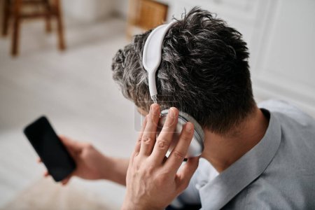 Un hombre trabaja desde casa mientras usa auriculares, sosteniendo un teléfono inteligente.