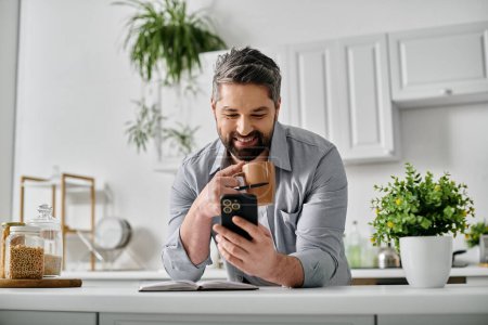 Ein bärtiger Mann in Freizeitkleidung sitzt an seinem Küchentisch und schaut auf sein Handy, während er eine Tasse Kaffee genießt..