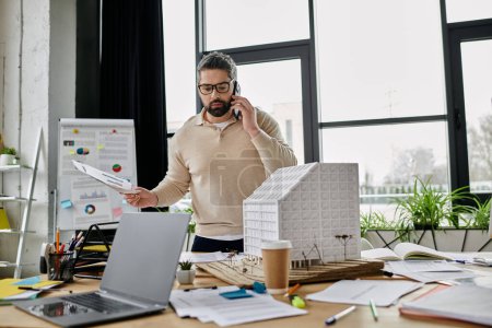 A handsome businessman with a beard works on building plans while on the phone in a modern office.