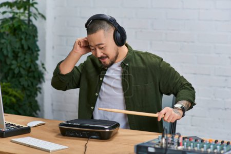 A young Asian man wearing headphones plays a drum pad in a recording studio.